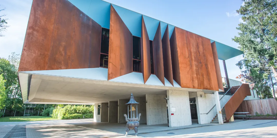 Exterior view of Wong Dai Sin Temple in Markham, featuring a modern structure with angled rust-coloured metal panels, a white elevated entrance supported by columns, and a small shrine beneath the overhang, surrounded by greenery.